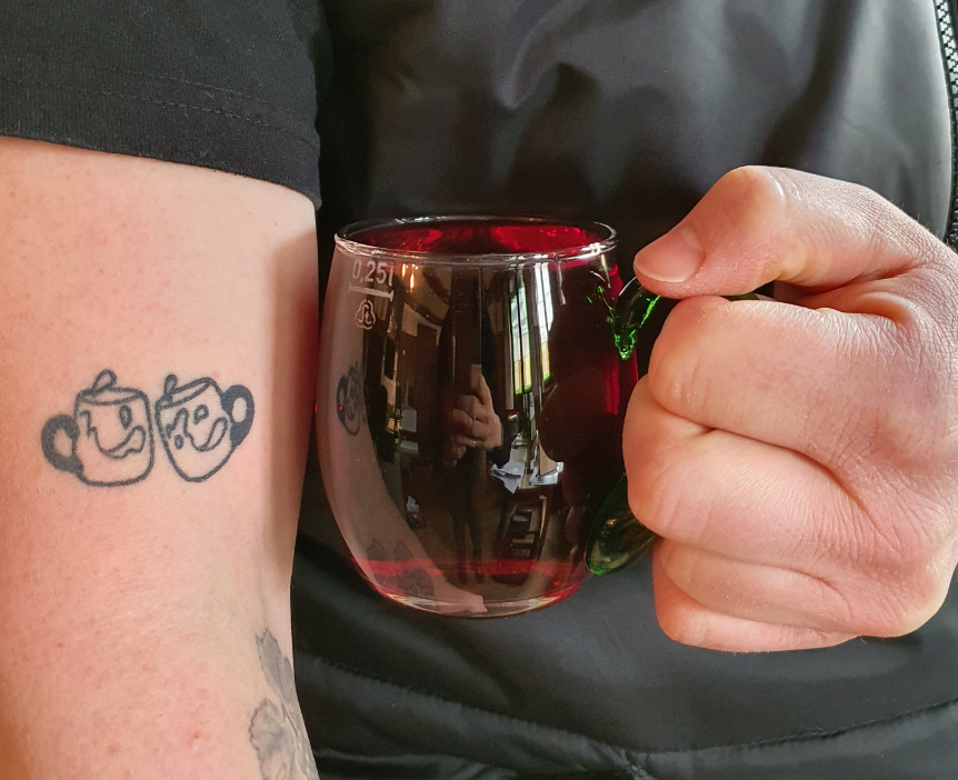 Man holding a mug of red wine next to his arm with a tattoo of clinking glasses