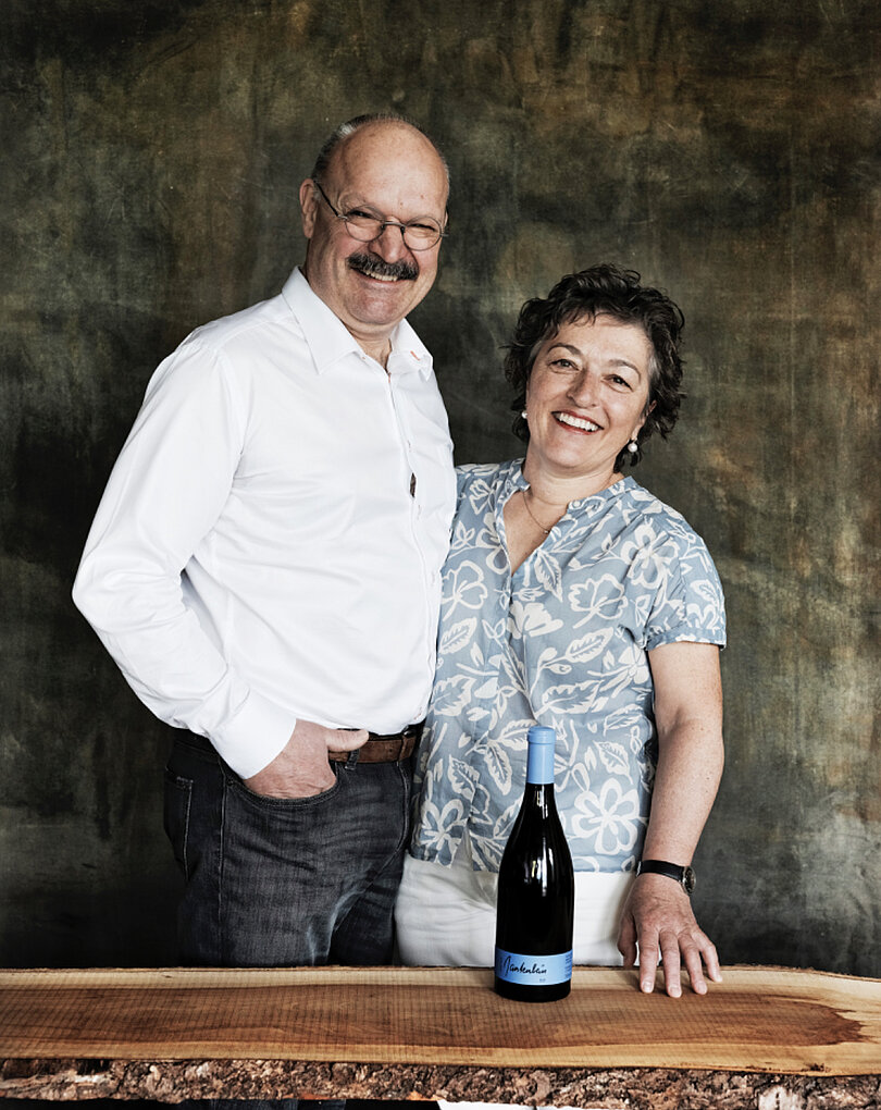 Portrait of Daniel and Marta Gantenbein smiling, standing behind a table with a bottle of their wine
