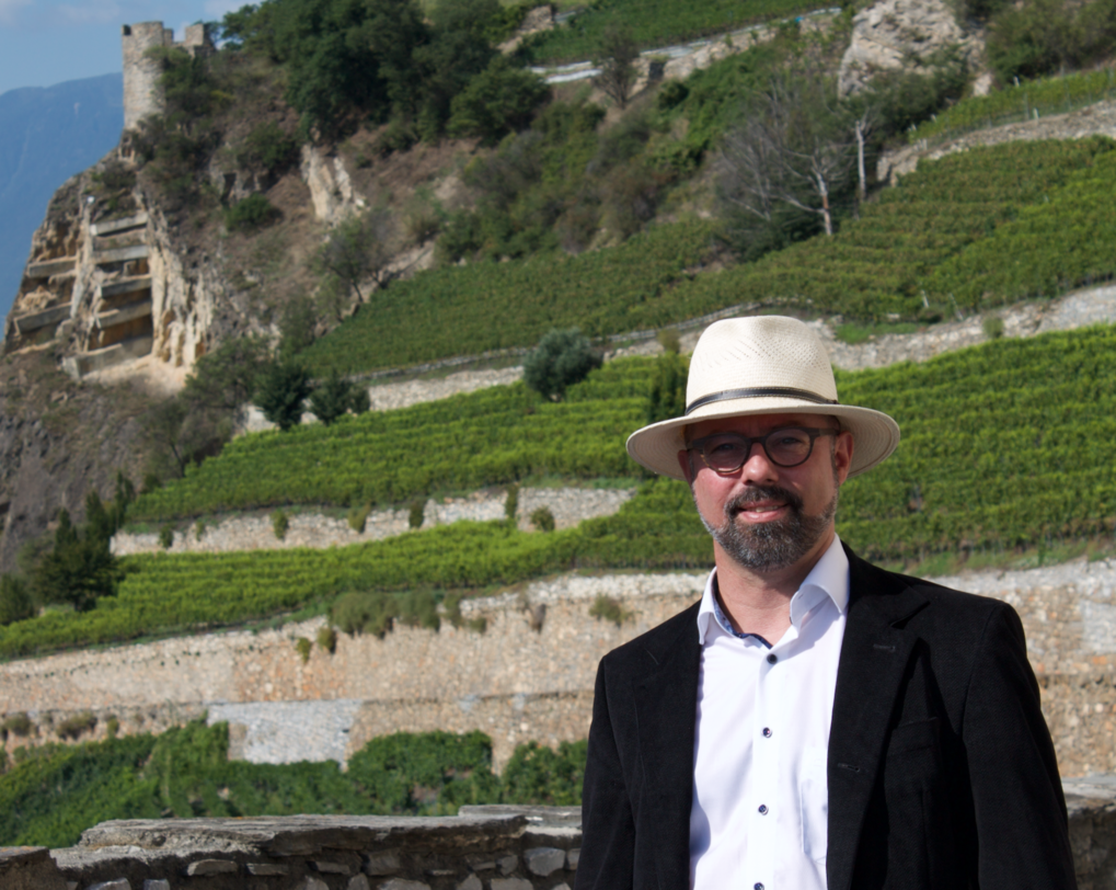 Photograph of Dr. Jose Vouillamoz dressed in a black blazer, white shirt and white hat stands in front of massive terraced vineyards with trees and cliffs in the far distance