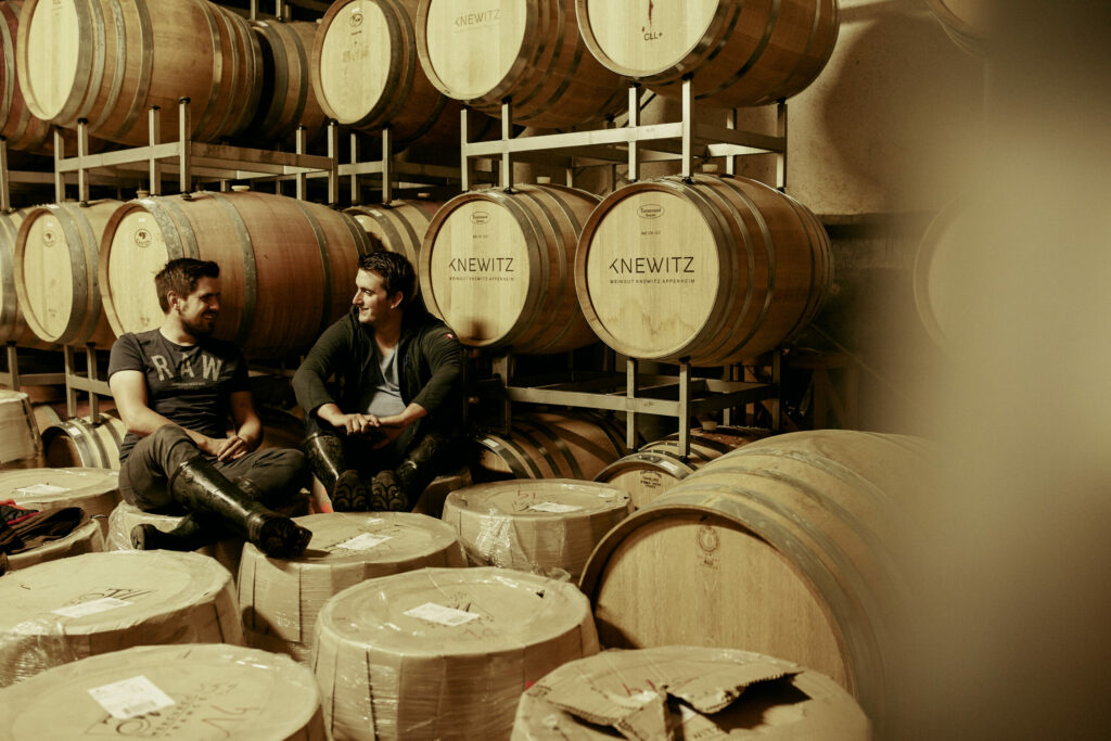 A photograph of Tobias Knewitz sitting on barrels of aging German Chardonnay in his Rheinhessen cellar