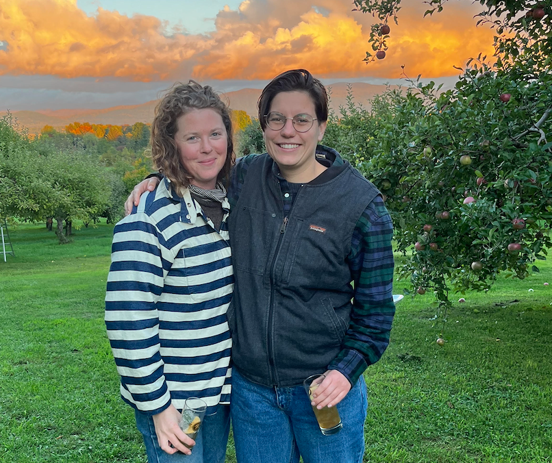 Owners of the queer wine shop Schmetterling stand in an orchard with mountains and sunset clouds behind them.
