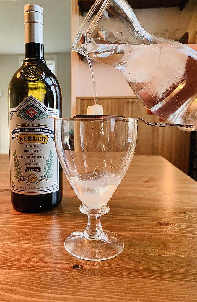 A photograph of a bottle of Kübler absinthe sits on a wood table, with an empty glass in the foreground into which water is being poured from a large glass decanter