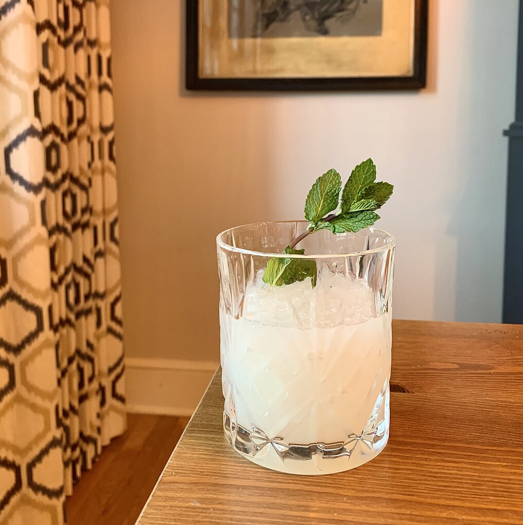 A photograph of a tumbler glass filled with icey, milky liquid and a mint spring sits on a wood table with a curtain and painting in the background