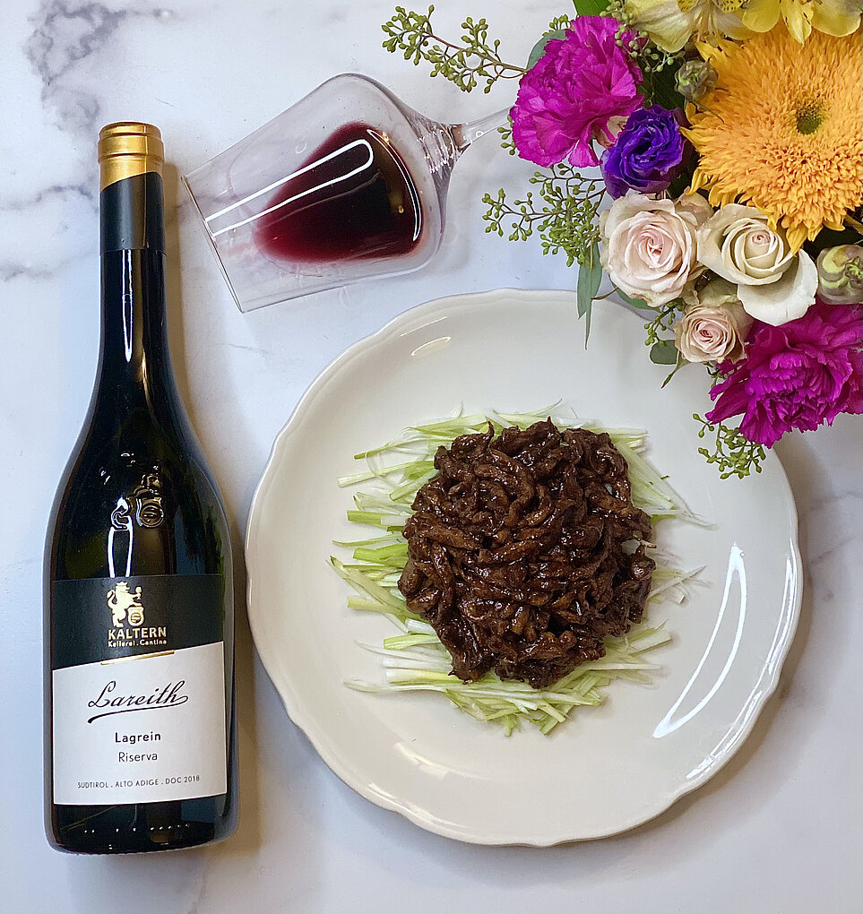 Bottle of Lagrein Riserva from Alto Adige next to a white plate of Jing Jian Rou Si beside a small bouquet