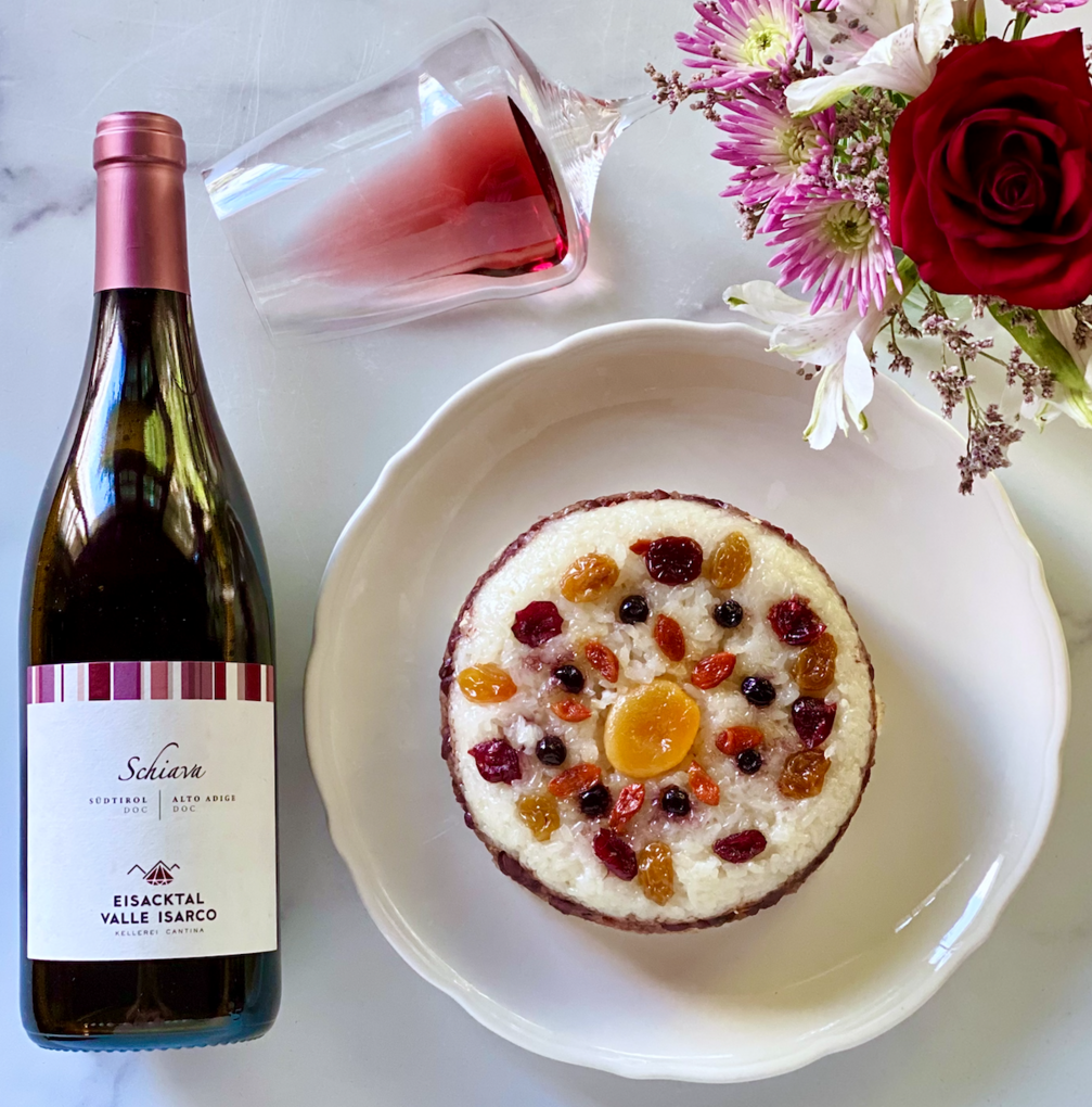 Bottle of red wine from Valle Isarco next to a white plate of Eight Treasure Rice on a white tablecloth bordered by a flower bouquet