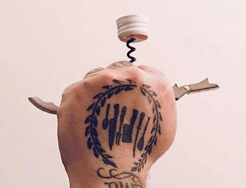A tattooed fist hold a corkscrew piercing a wine bottle screwcap.