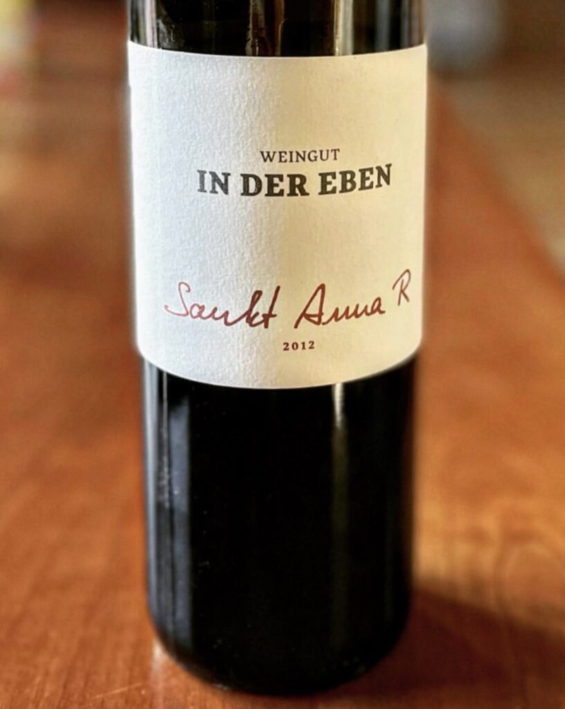 A dark glass wine bottle stands on a wooden table. The white label features black lettering reading Weingut IN DER EBEN and in red script Sankt Anna R 2012