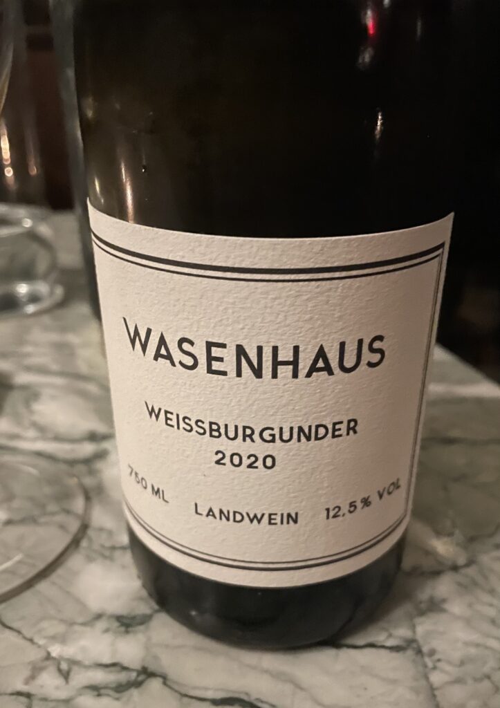 Against a marble table top background, a dark glass wine bottle with a minimalist white and black wine label ready WASENHAUS WEISSBURGUNDER 2020