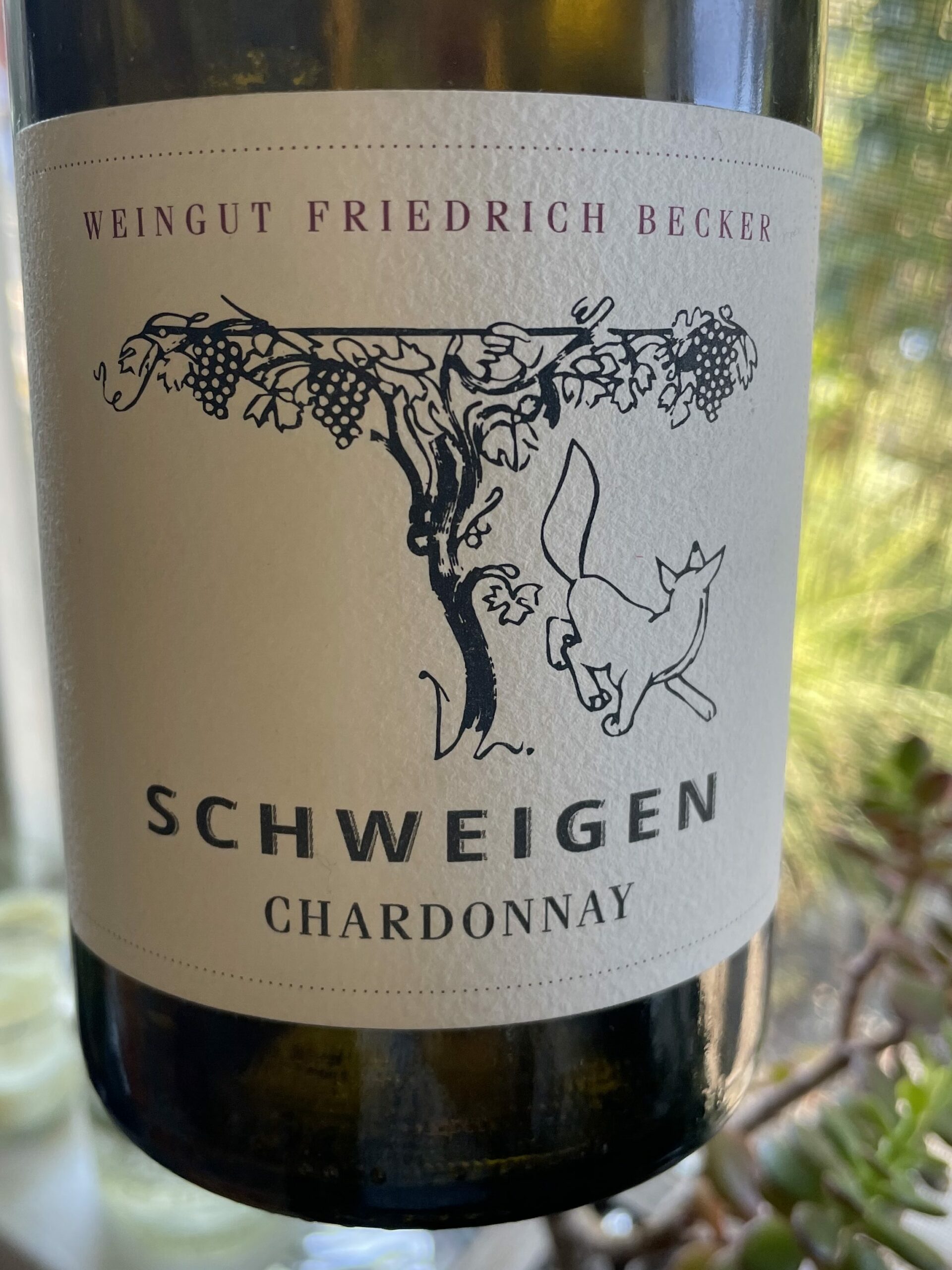 Wine botle in foreground with white label and black lettering featuring logo and SCHWEIGEN.