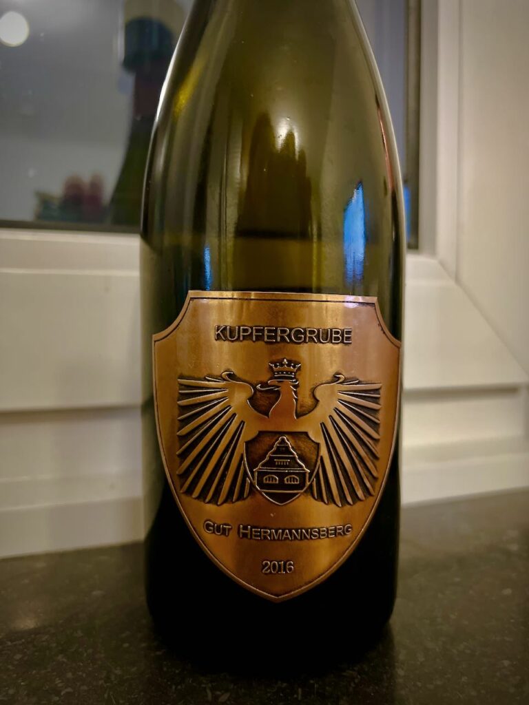 Against a window frame, a wide dark glass wine bottle with a copper label, spread-wing eagle and black lettering reads KUPFERGRUBE GUT HERMANNSBERG 2016