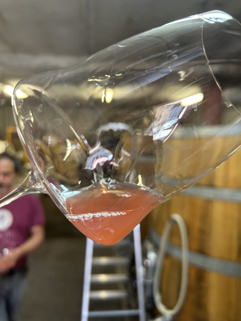 A glass of wine in the foreground, with barrels and winemaker in the background.