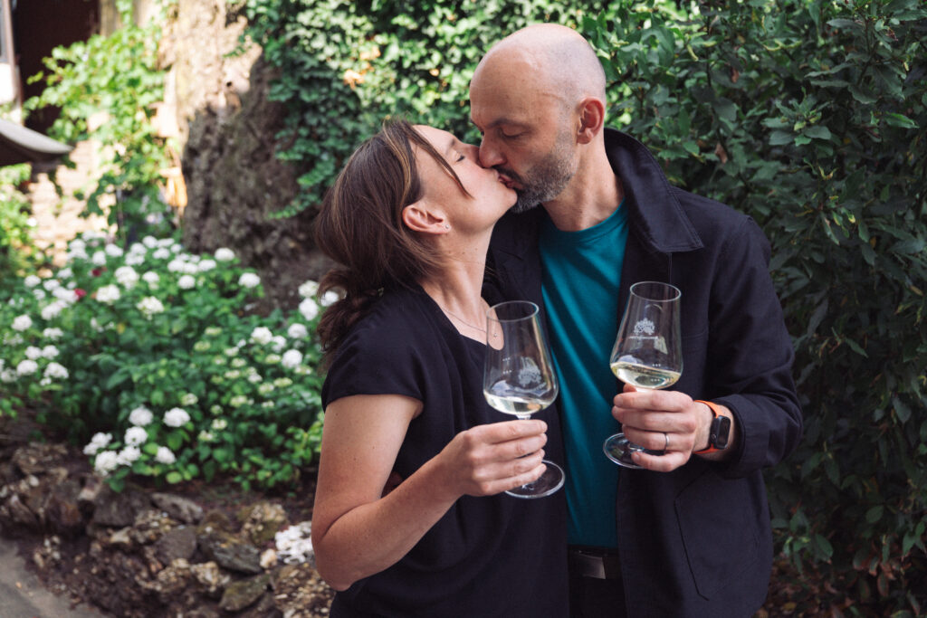 Caroline and Sylvain Diel kiss while holding two wine glasses in the garden of their Nahe winegrowing estate