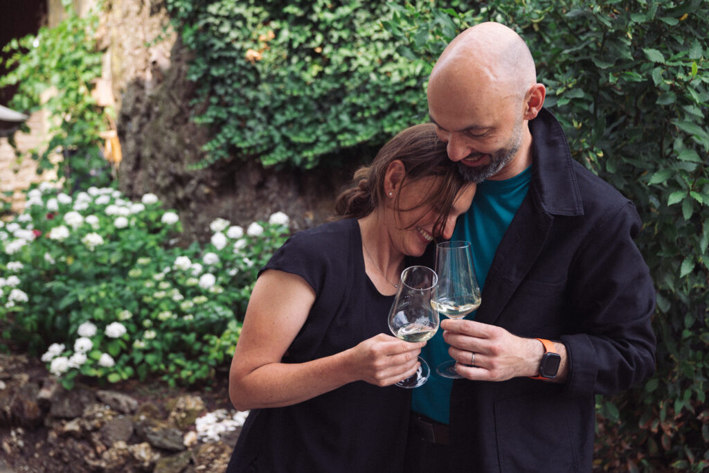 Caroline and Sylvain Diel lean into each other holding two wine glasses in the garden of their Nahe winegrowing estate