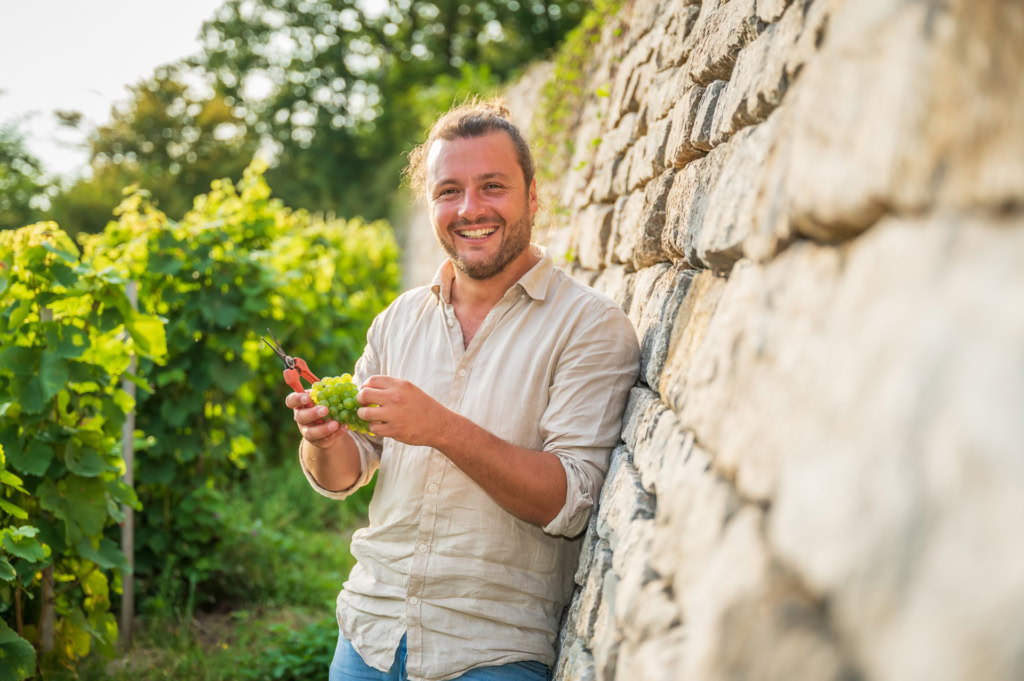 Nico Olinger smiles and stands against a light stone wall in vineyards