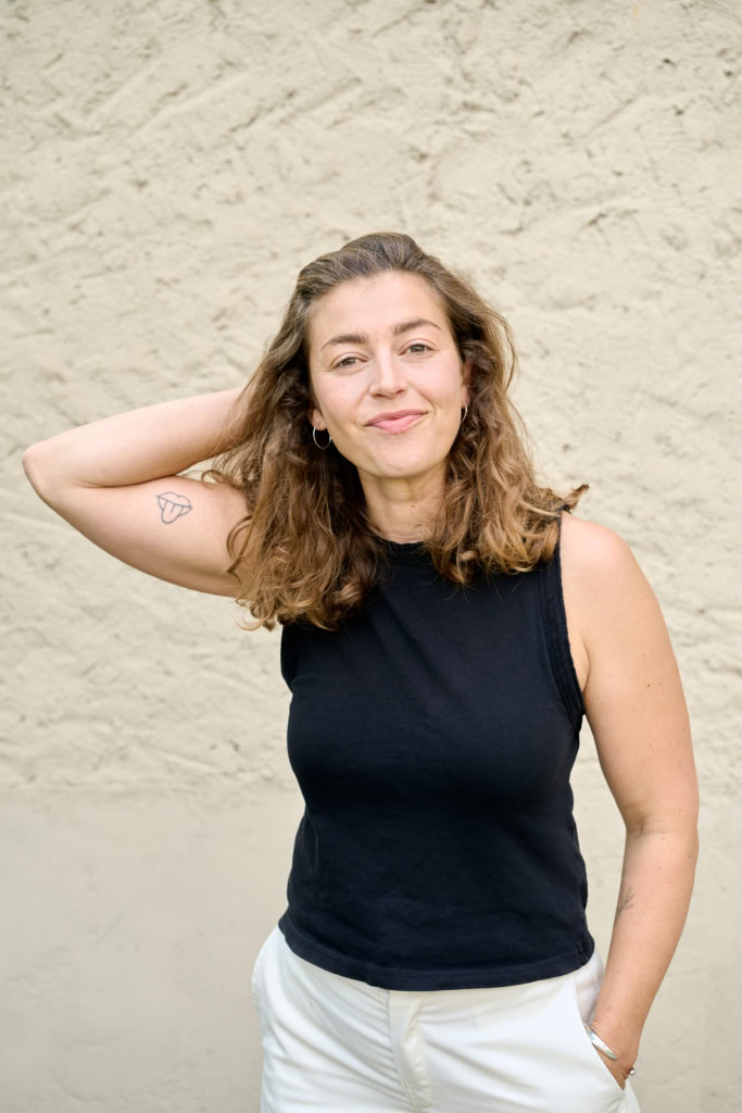 Franziska Schömig stands in a black sleevelss top against a white wall, with a smile and one arm at her side, the other behind her head.