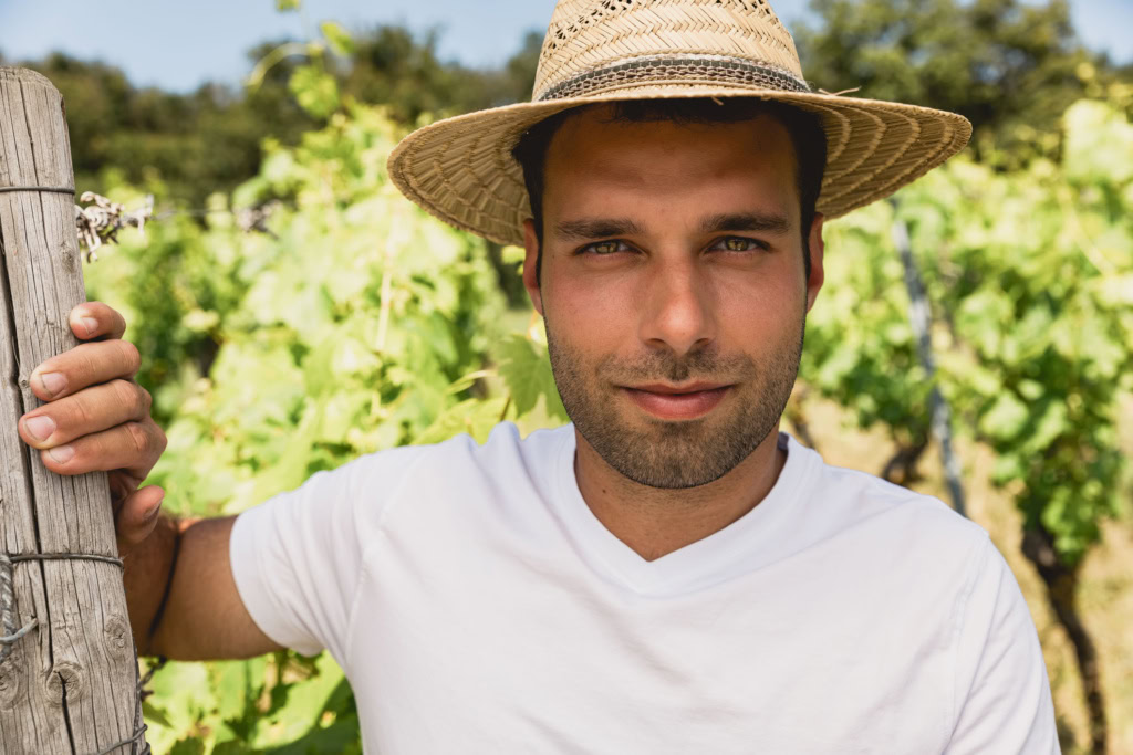 Close-up of Franken wine grower Peter Leipold in a white t-shirt and straw hat standing among vineyards in summer.