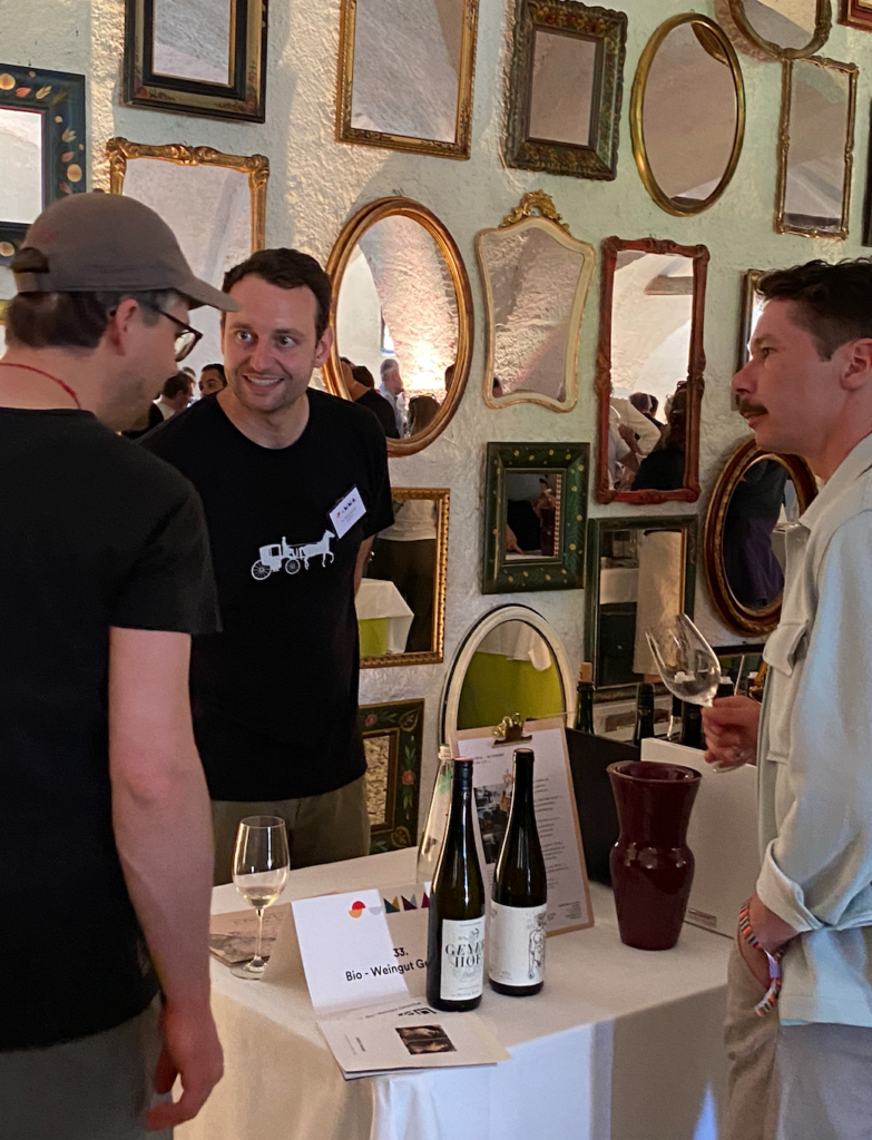 Three men lean into a conversation over wine bottles against a mirror-lined wall.