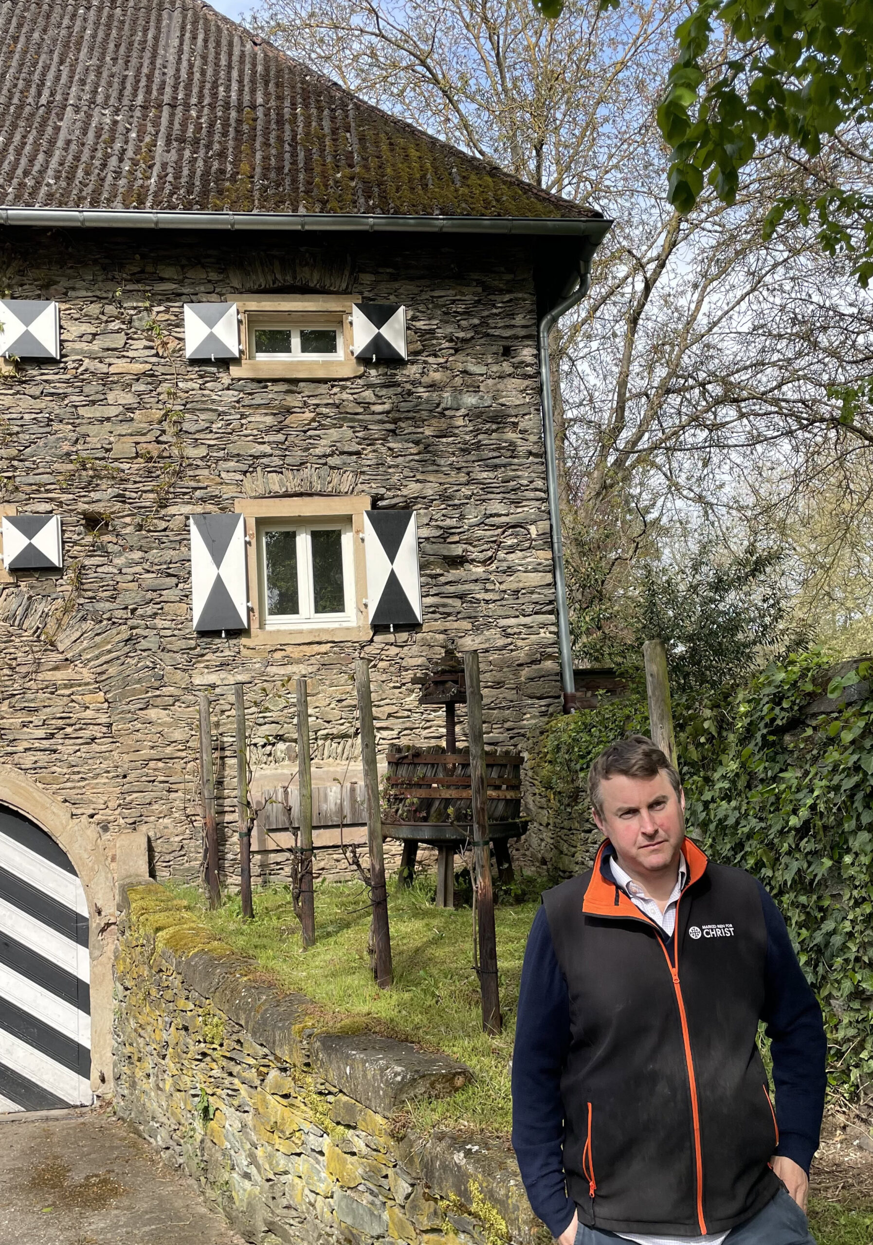 Felix Prinz Salm stands outdoors in front of his ancient stone winery decorated with black and white-painted shutters and door