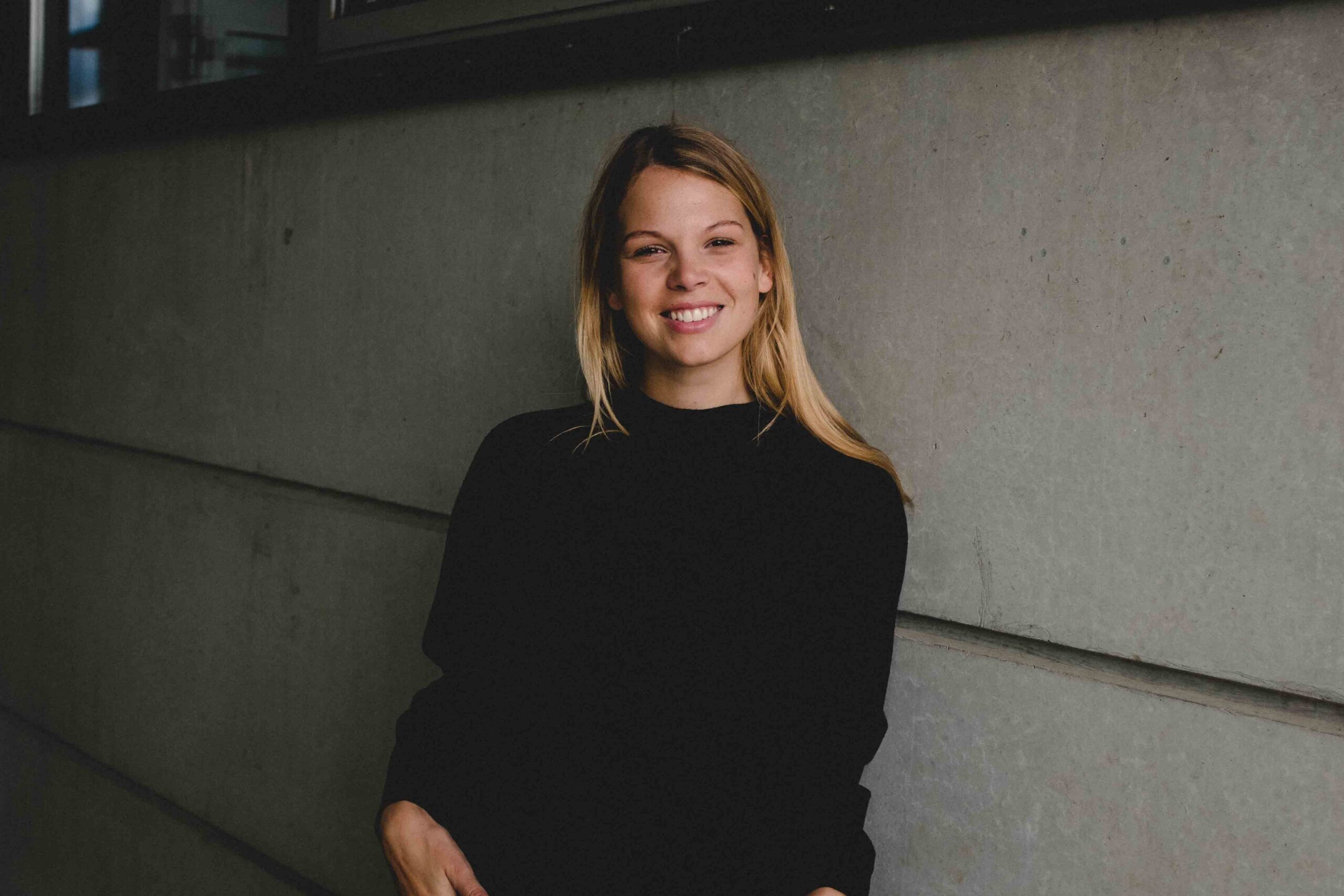 Theresa Olkus stands in a black shirt against a white wall, smiling.