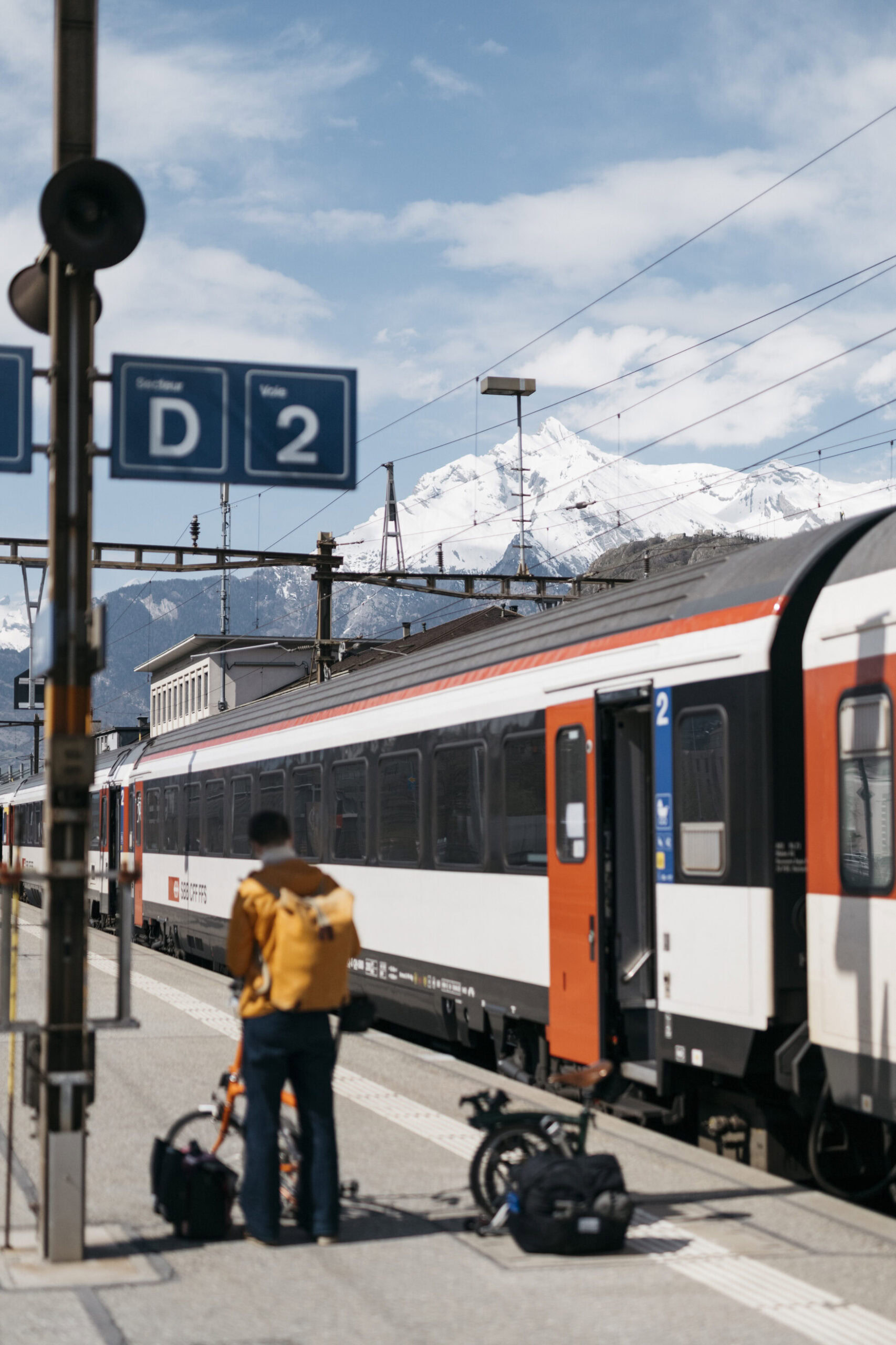 A colorfully dressed woman stands on a train platform with a Swiss train and the snow-covered Alps in the background