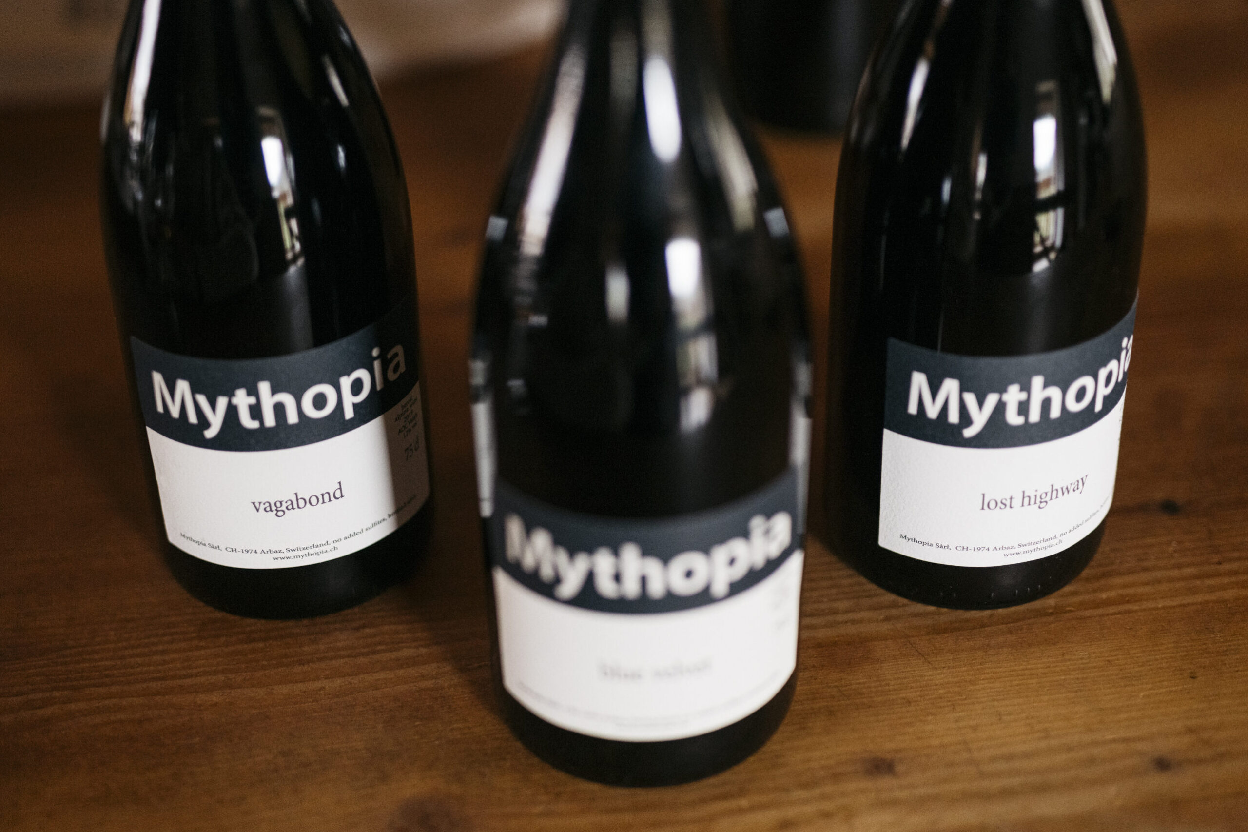 Three bottles of Mythopia Pinot Noir stand on a wooden surface.