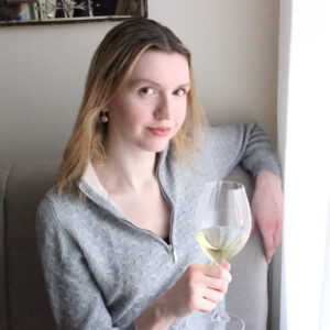 Wine writer Alexandra McInnis sits on a chair with a glass of white wine in her hand next to a window with a translucent curtain.