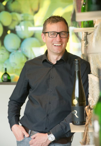 Franken winemaker in black rimmed glasses leans against a limestone wall while smiling surrounded by his wine bottles