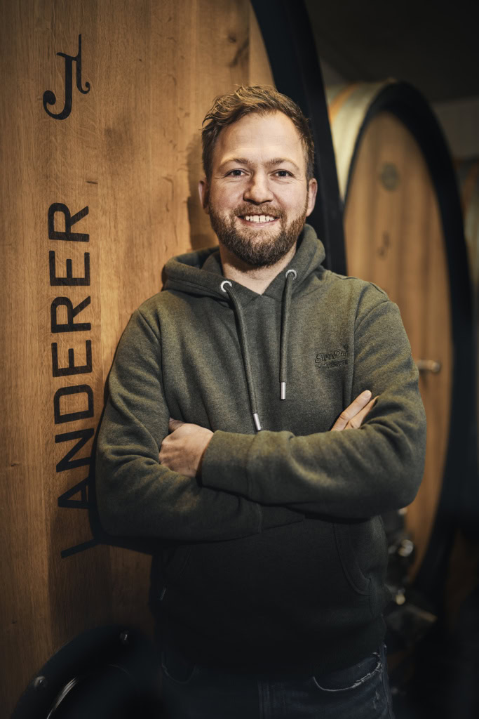 Johannes Landerer stands smiling with his arms folded in front of a large wooden barrel. 