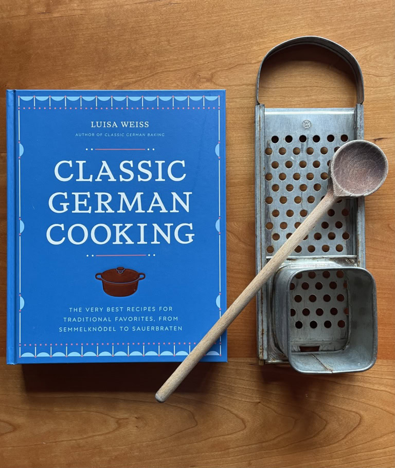 A blue hardcover book with the title Classic German Cooking by Luisa Weiss rests on a wooden table next to cooking implements.