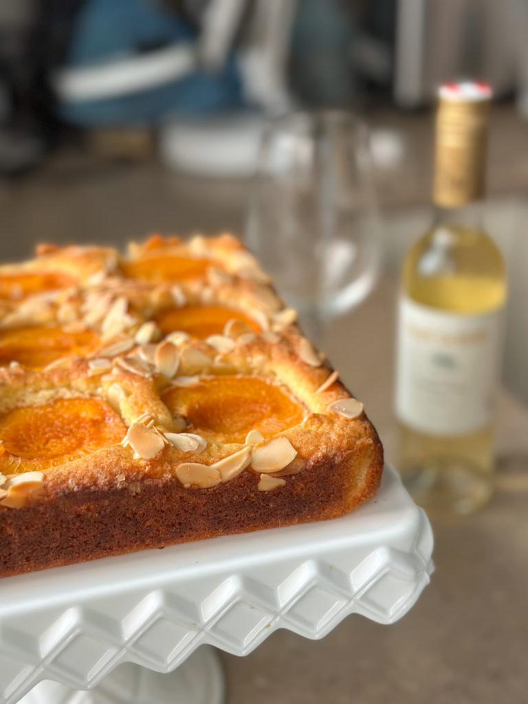 A cake with almonds on top on a white plate with a bottle of golden-colored wine and a glass in the background