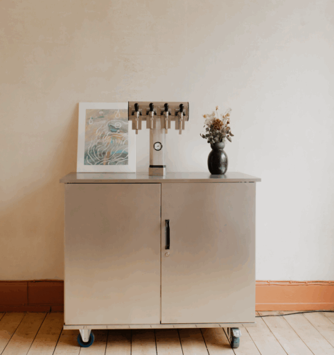 A stainless steel cabinet sits in an other wise empty space. Taps for kegged wine, a painting, and a vase of flowers sit on top.