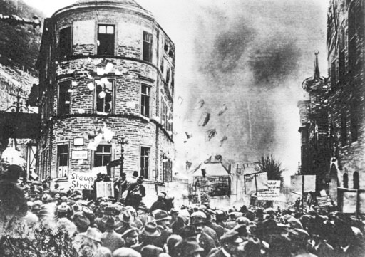 Grainy black and white photo of uproar in the streets, filled with people, a vineyard detectable faintly in the background. A sign reads "Steuer Streik".